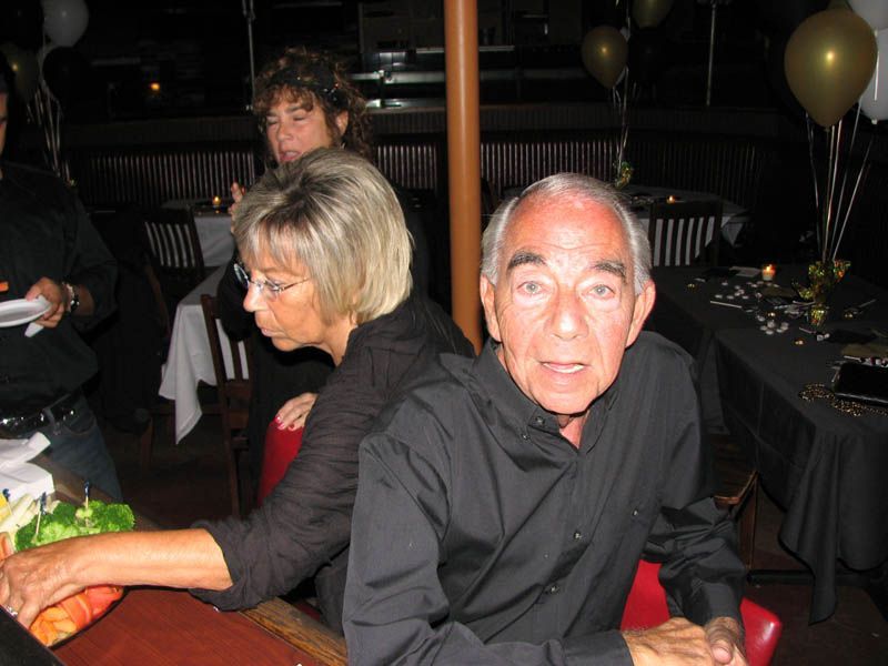 Man in black shirt at a party, looking at the camera. Others sit nearby, tables with food, balloons in background.