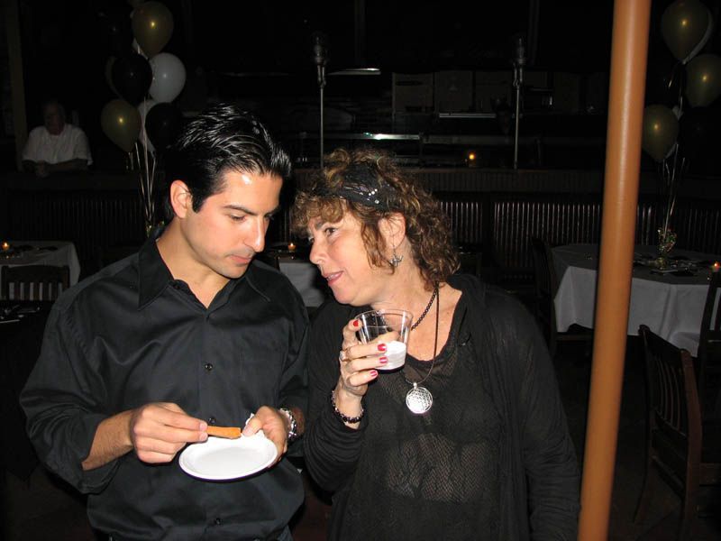 Man and woman at an event, man holds a snack, woman holds a drink, indoors with balloons.