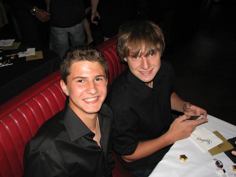 Two young men smiling, sitting at a table with white tablecloth and red booth seating, likely at a restaurant.