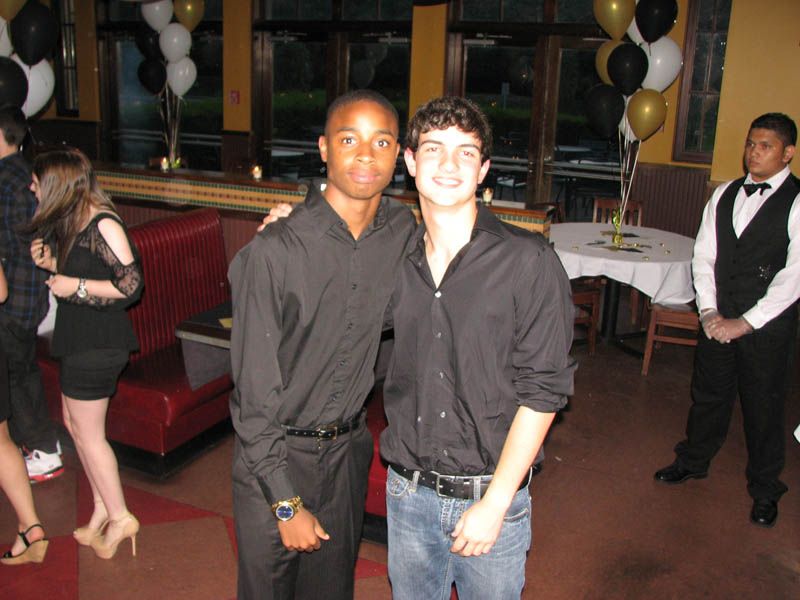 Two young men in black shirts pose at a formal event, with decor and others visible.