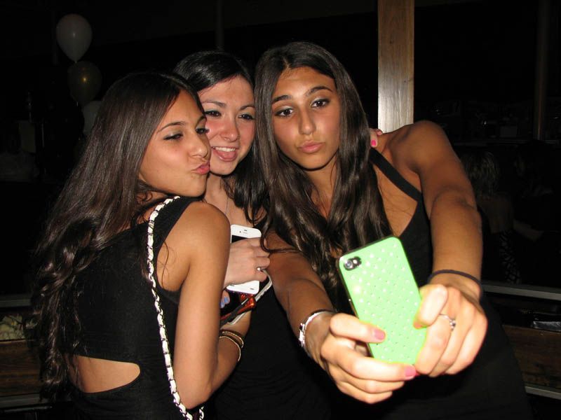 Three people taking a selfie in a dark setting, one holding a green phone, all with dark hair and wearing black dresses.