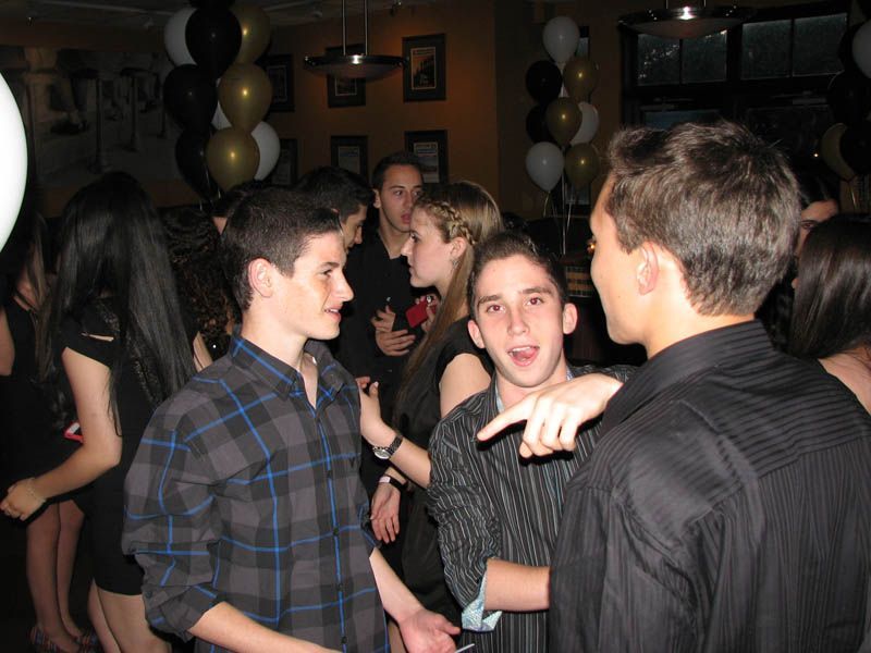 People at a party, some talking and gesturing. Black and gold balloons, dark setting.