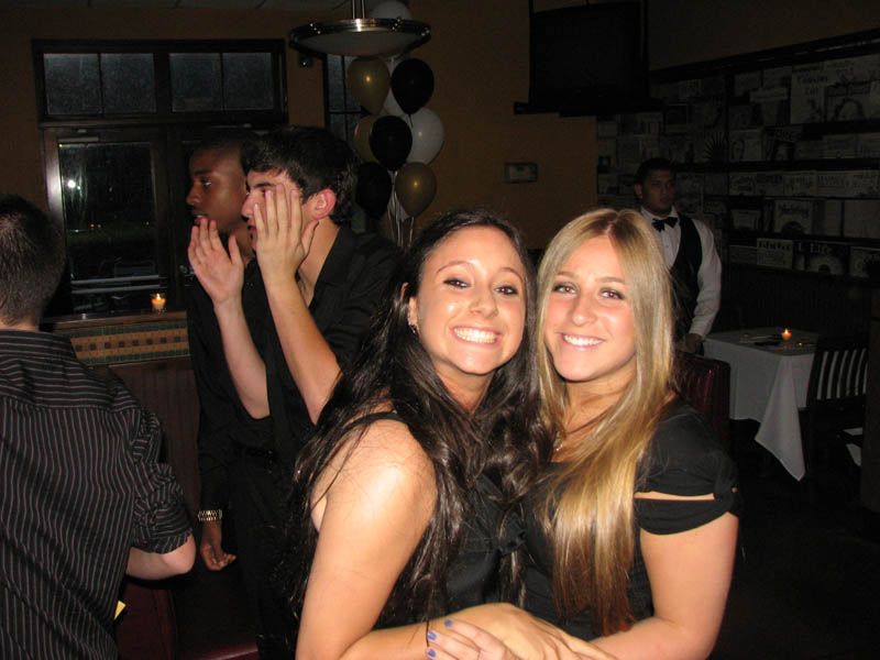 Two smiling women posing, others in dark room with balloons, waiter in background.