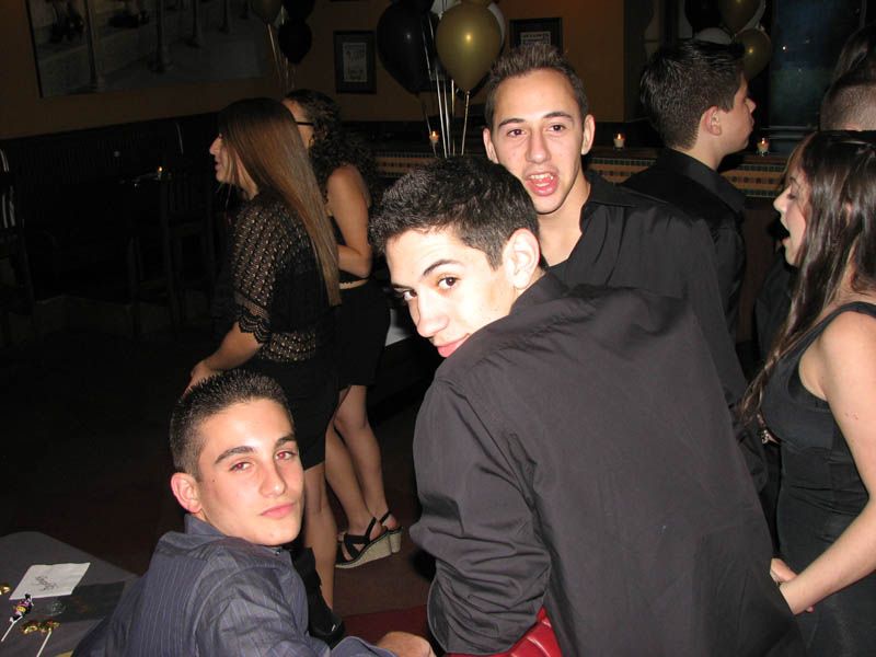Group of teens at a party. Some are looking at the camera, others in the background. Black attire, balloons, interior.