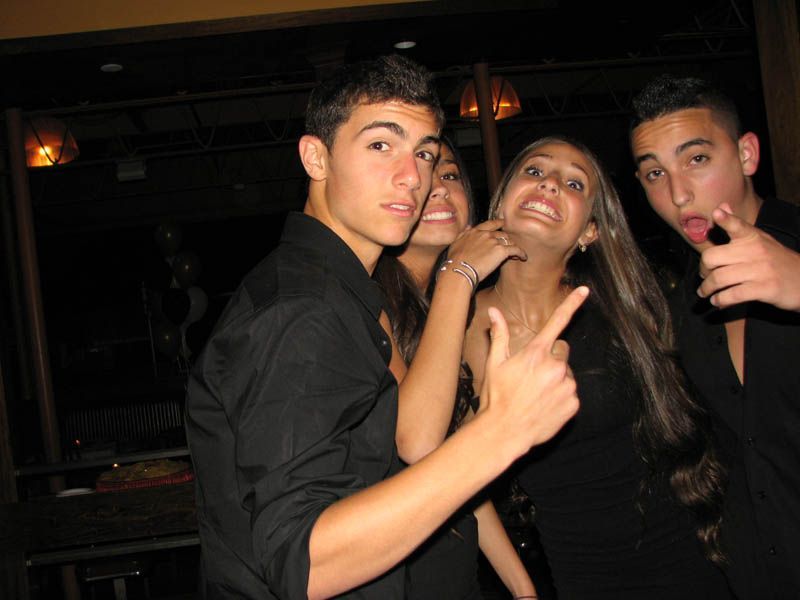 Four young people in a dark room; two men, and two women. They're smiling and pointing towards the camera.