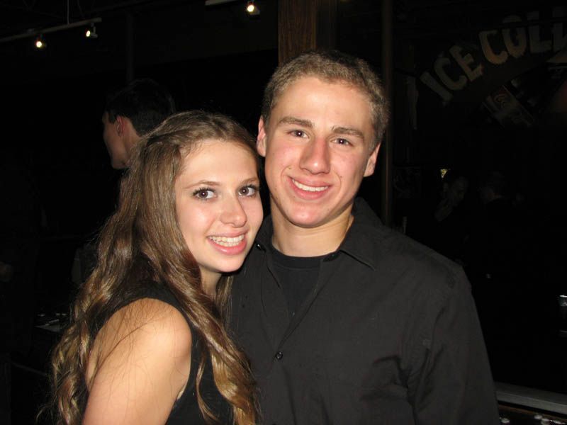 A smiling couple poses close together indoors. The woman has long brown hair and the man wears a black shirt.