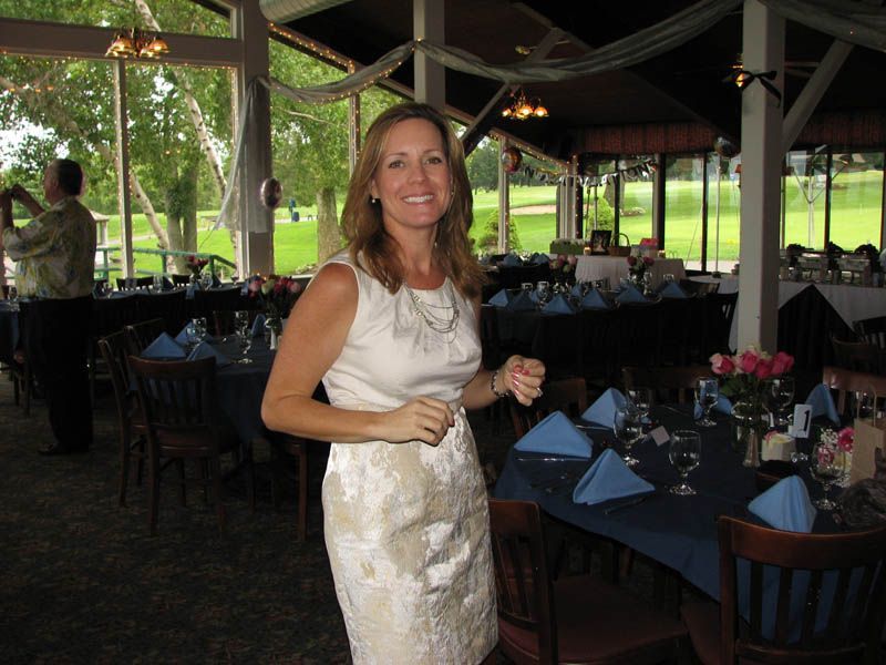 Woman in white dress smiles, stands near tables set for event in a venue with outdoor view.