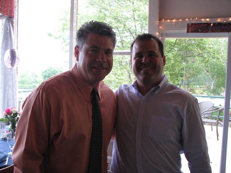 Two men smiling, one in an orange shirt and tie, the other in a light blue shirt, near a window.