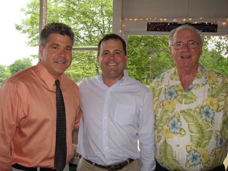 Three men, arm-in-arm, smiling. One in orange shirt and tie, middle one in light blue shirt, right one in floral shirt.