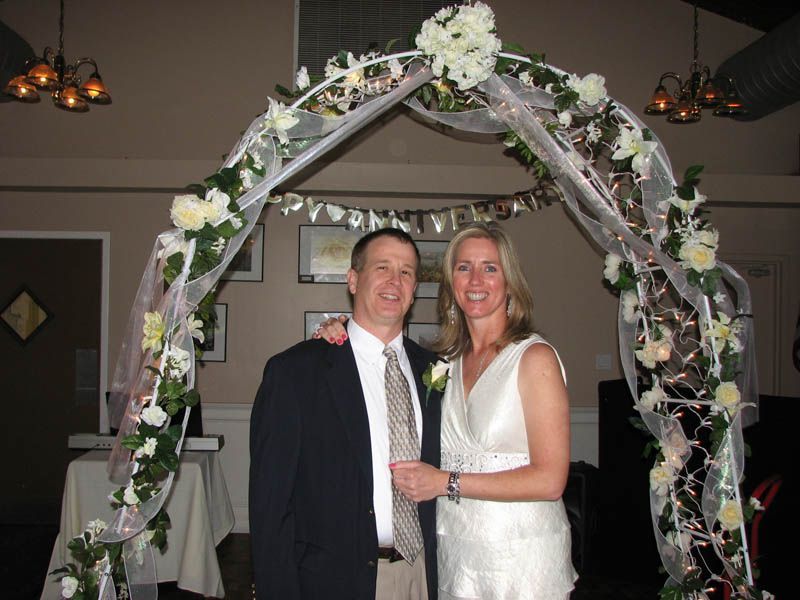 Couple in wedding attire under a floral arch. Man in suit, woman in white dress, smiling. Indoor setting.