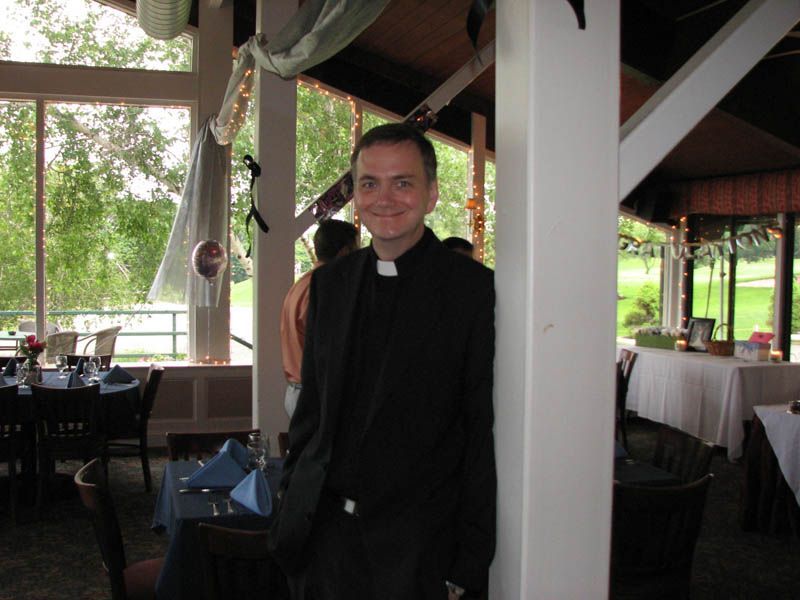 A man in a clerical collar stands in a dining area, leaning against a white pillar.
