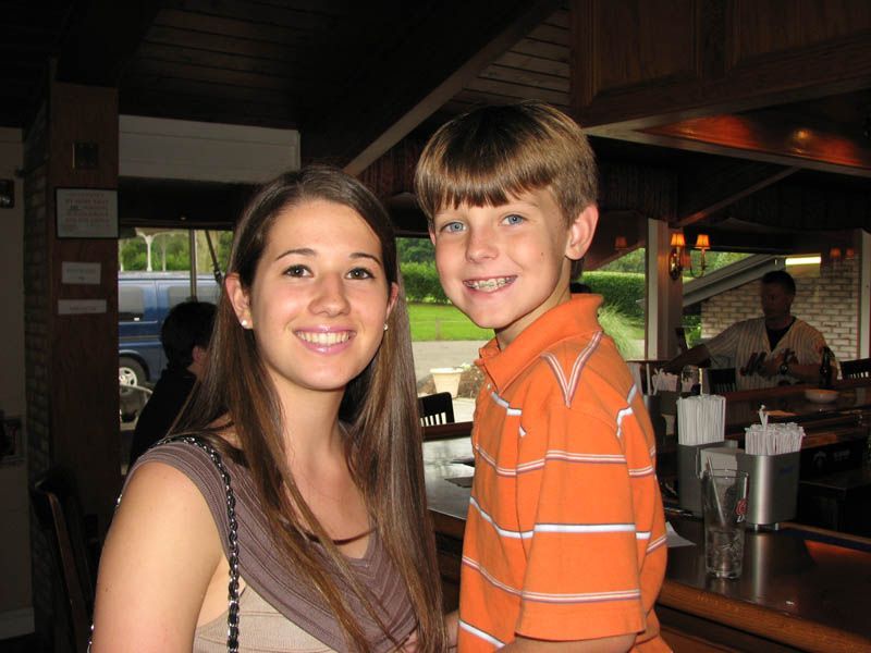 Woman and boy smiling, indoors near a bar; woman with brown hair, boy in orange shirt.