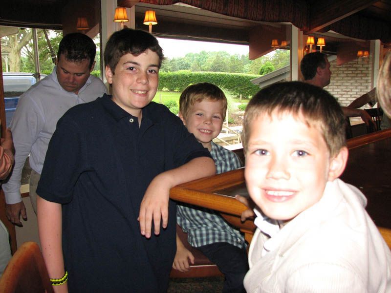 Three smiling boys at a bar; one in front with fair hair, others behind. Man in background.