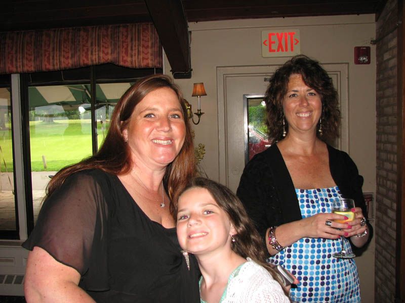 Three smiling people posing indoors: two women and a child. One woman holds a drink.