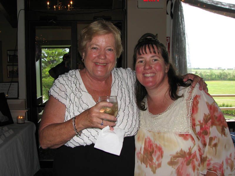 Two women smiling, arm-in-arm; one holds a drink. Window in background.