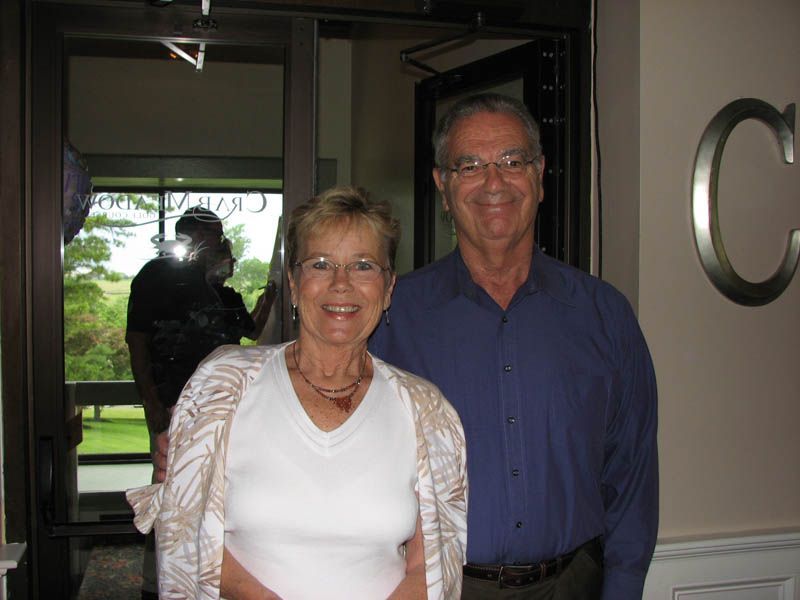 Smiling couple standing in front of a glass door; man in blue shirt, woman in white top with shawl.