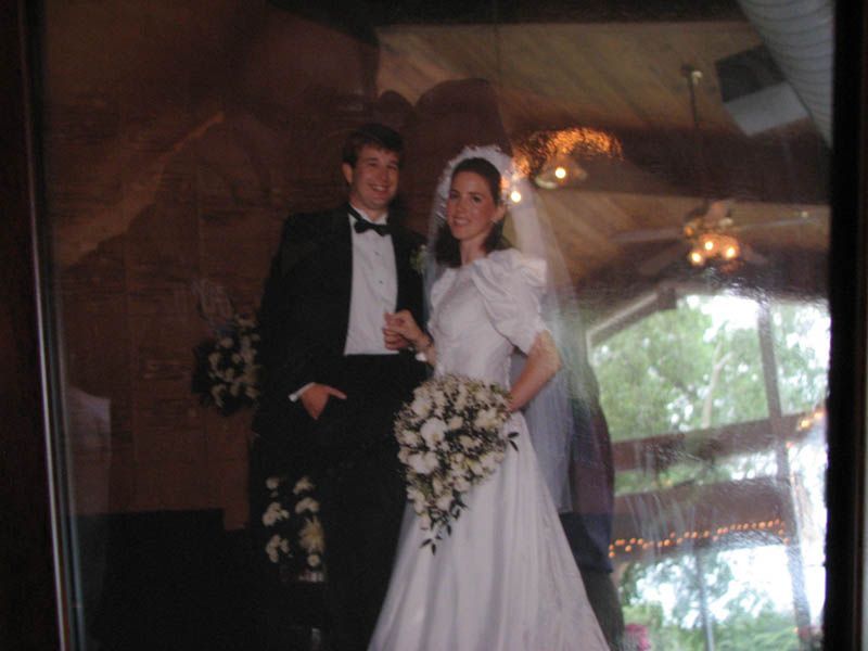 Couple in wedding attire; woman holds bouquet. Man in tuxedo, woman in white dress and veil, standing indoors.