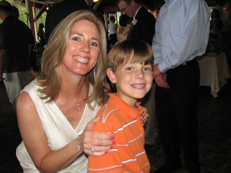 Woman in white dress smiles, arm around boy in orange shirt; both smiling. People in background, indoors.