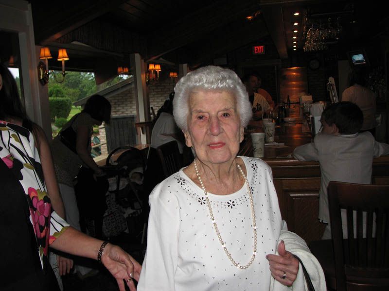 Elderly woman at a restaurant, wearing white top and pearl necklace, smiling.