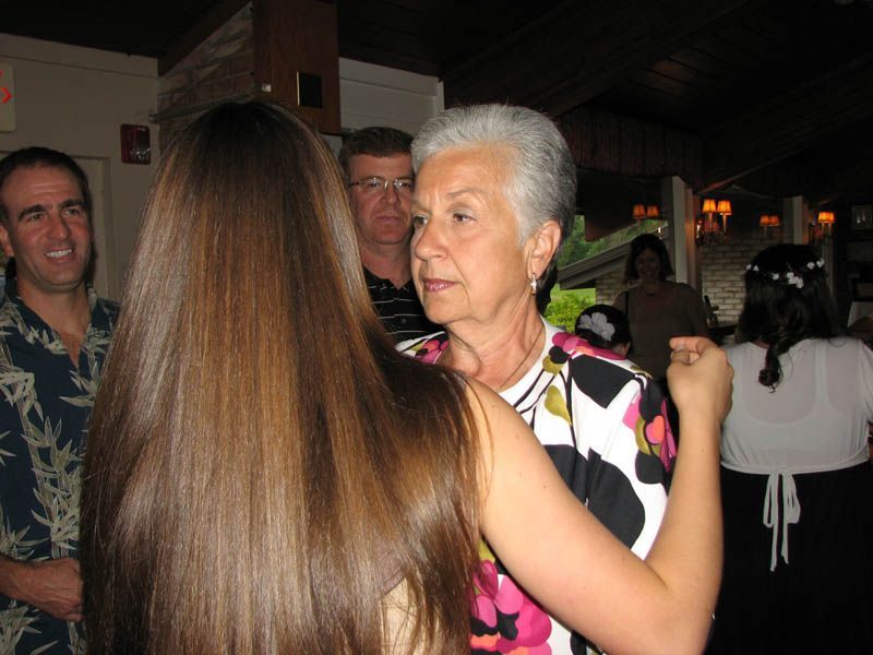Woman with gray hair dances with another woman. People watch nearby at a social event.