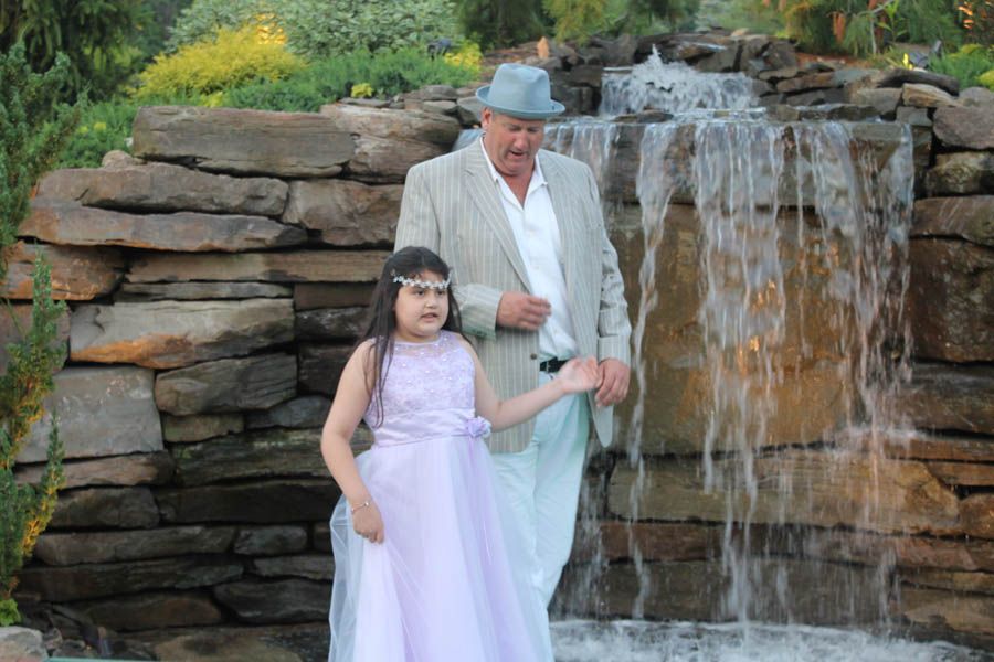 Man and girl in front of small waterfall; girl in lavender dress, man in tan jacket and hat.