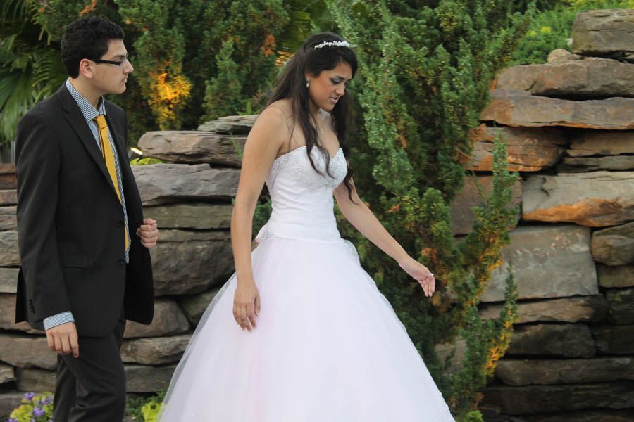 Bride in white dress walks outdoors, groom in suit follows. Stone wall and greenery in background.