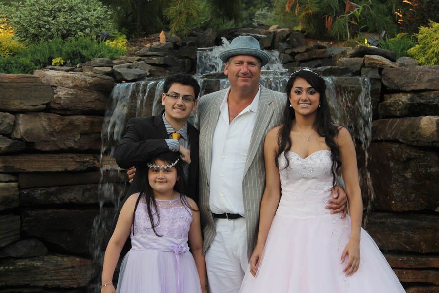 Family posing in front of a waterfall: older man with fedora, two young adults, and a child in formal wear.