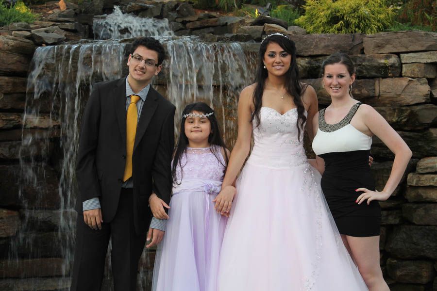 Four people pose in front of a small waterfall. Woman in white gown, man in suit, girl in lilac dress, woman in black dress.