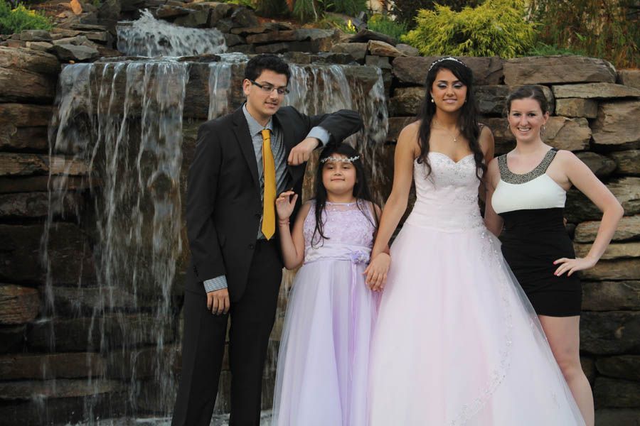 Four people pose in front of a small waterfall. Woman in white gown, man in suit, girl in lilac, woman in black dress.