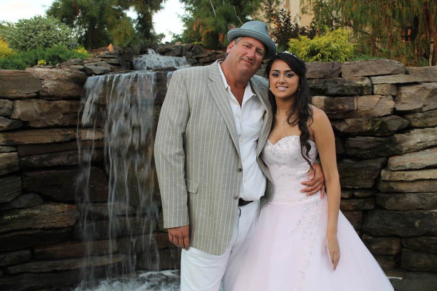 Man in a light-colored suit and woman in a pink gown pose by a small waterfall.