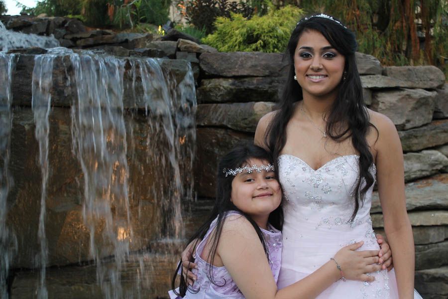 Two smiling people pose by a small waterfall. One wears a strapless white dress, the other a lavender dress.