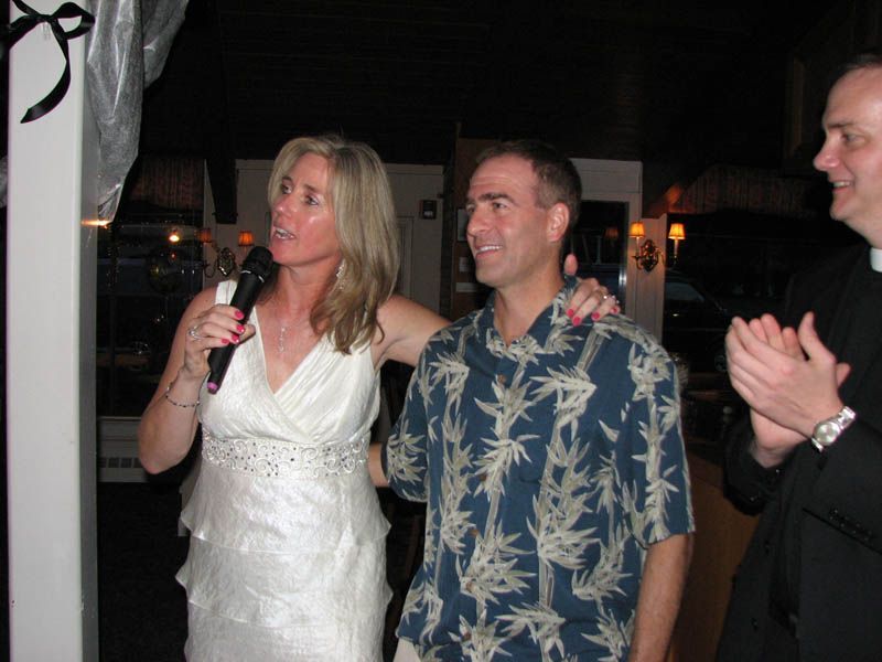 Woman in white dress speaks into a microphone, man in blue shirt beside her, priest clapping.