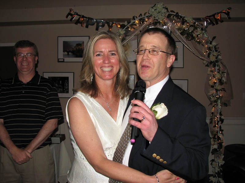 Woman and man smiling, man holding microphone, near decorative arch and man in striped shirt.