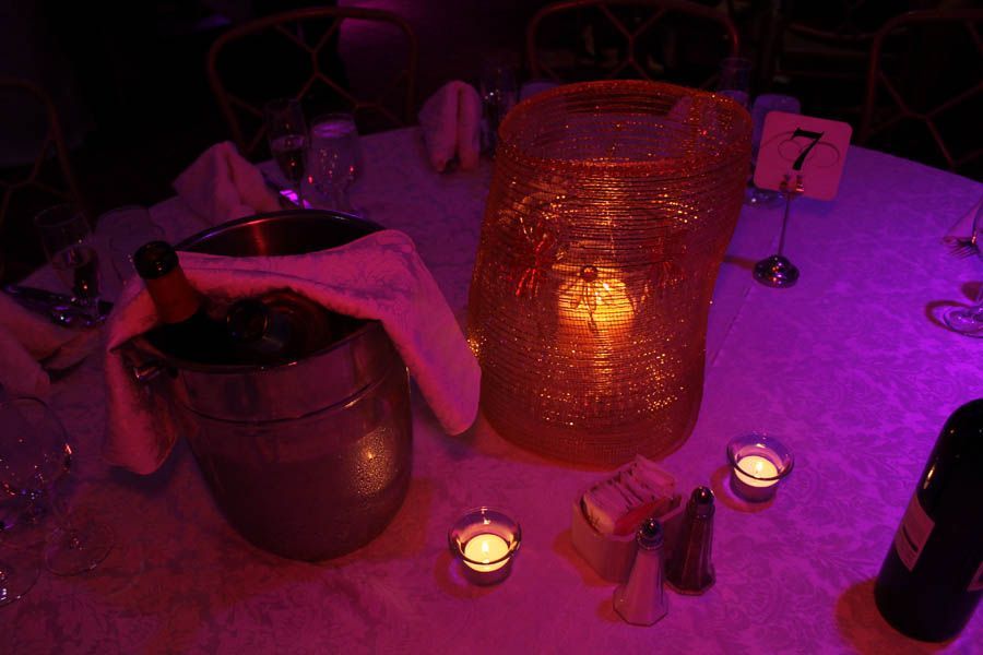 Table setting with bucket, bottle, tall orange glass, candles, and table number 7 under pink lighting.