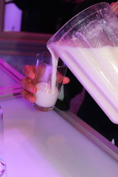 A hand pours a thick, white liquid from a blender into a clear glass, on a pink-lit surface.
