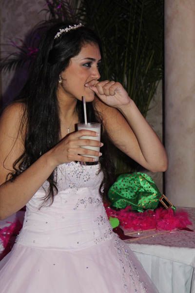 Woman in a pink dress, tiara, holding a drink with a straw, biting her nail. Party setting.