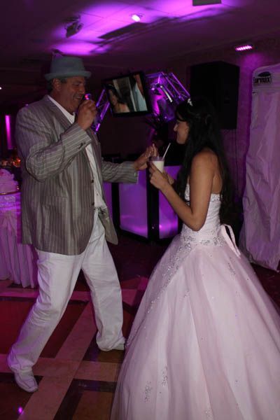 Man in a suit and hat dances with a woman in a wedding dress, both holding straws to a drink, indoors.
