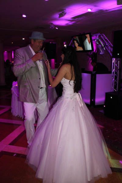 A woman in a white wedding dress dances with a man in a light gray suit and hat at a reception.