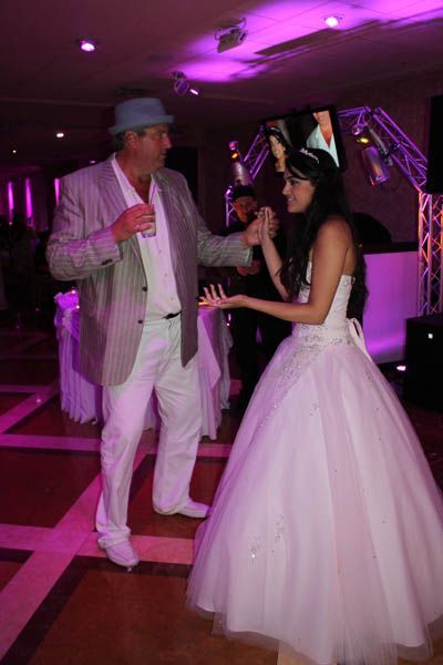 Man in light suit dances with a woman in a white ballgown, under purple lights.