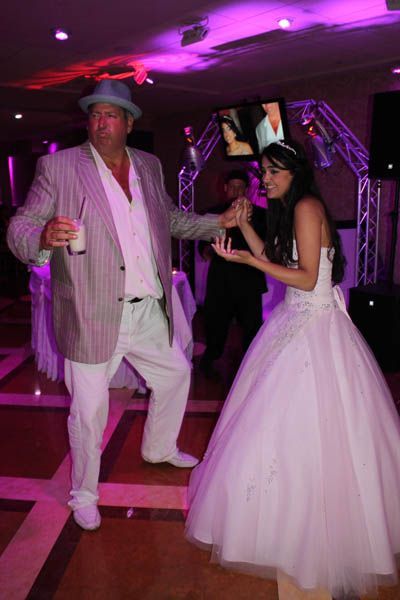 Man in white suit dances with woman in white gown. They are in a room with purple lighting.