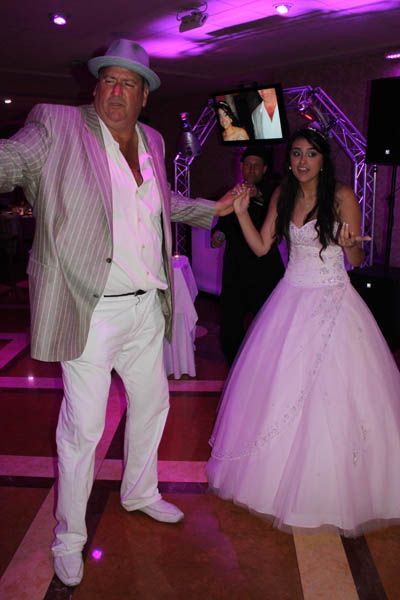 Man in white suit and hat dancing with a girl in a white gown at a party with pink lighting.