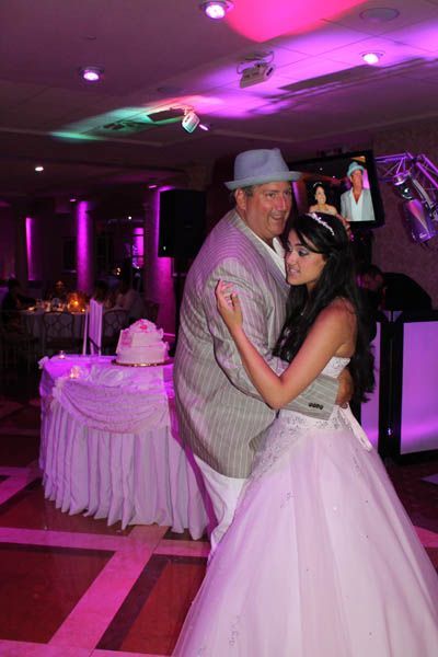 Man in suit and hat dances with woman in white gown at event. Cake and decorations in background.