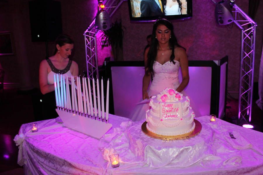 A young woman in a white dress stands by a tiered cake, facing a stand of candles on a table in a dimly lit room.