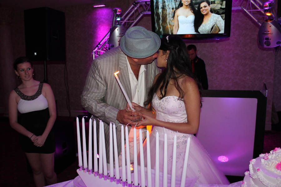 Couple kissing while lighting candles on a cake at a party, with a screen and DJ booth in the background.