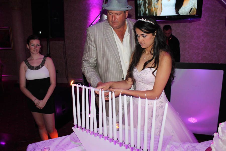 Girl in gown and man in suit light candles on a tiered cake; a woman watches nearby.