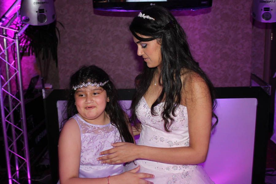 Two women, one in a white dress, one in a lavender dress, stand together indoors with purple lighting.