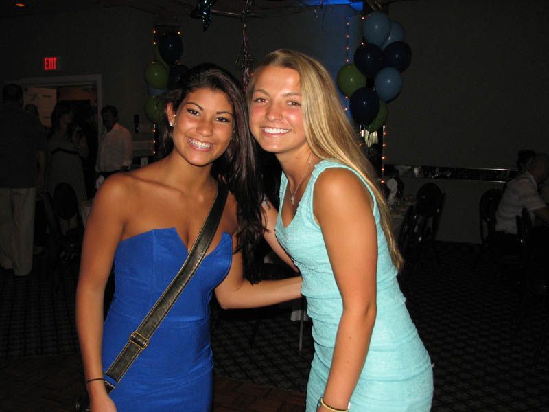 Two women smiling, posing at an event. One in a blue strapless dress, other in a light blue dress. Balloons in background.