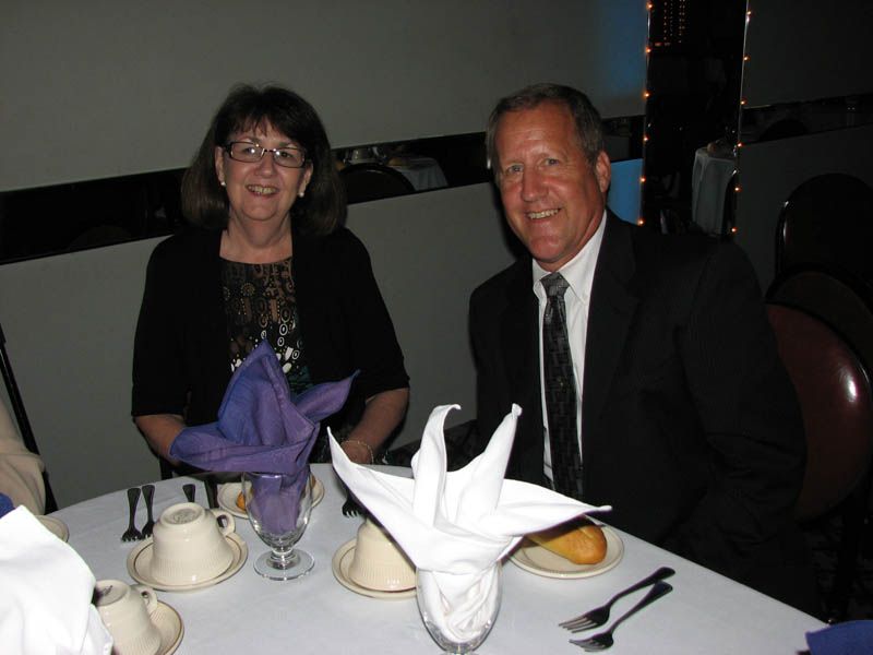 Couple at a formal table, woman in black blazer, man in suit. White tablecloth with napkin art and tableware.