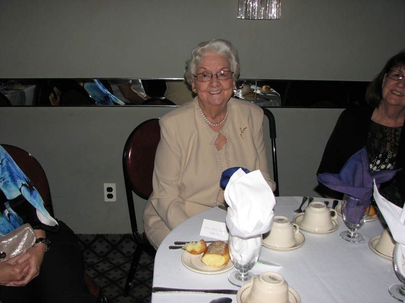 Elderly woman smiling at a table, wearing a beige suit, seated at a formal event.