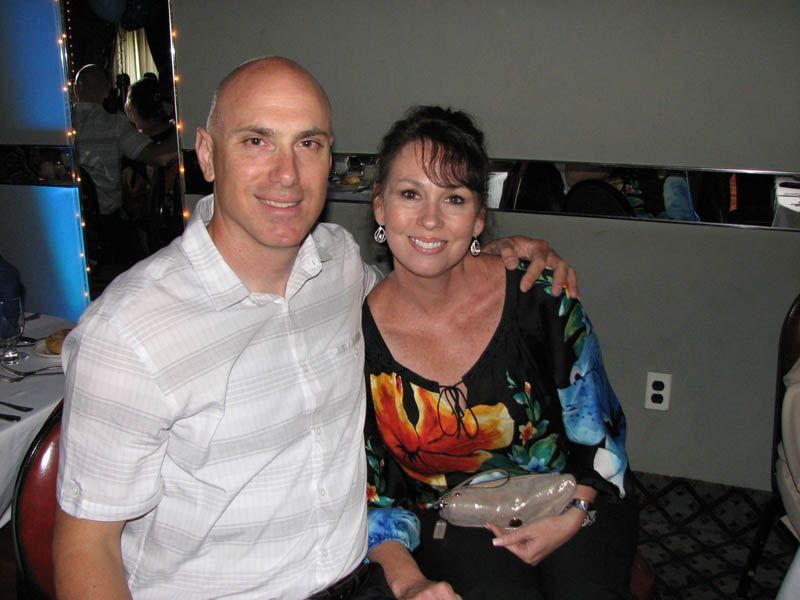 Man and woman smiling, indoors. Man with shaved head in white shirt, woman with dark hair in floral top, arm around him.